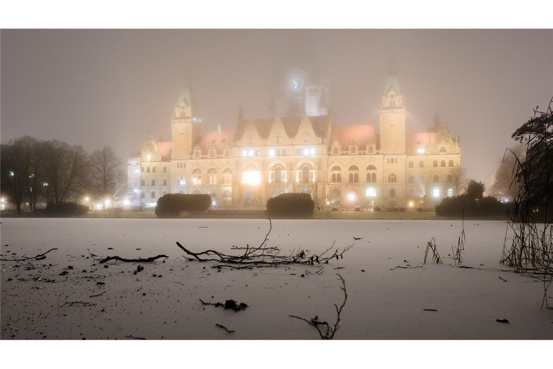 Das Neue Rathaus wird am frühen Morgen bei Nebel und Temperaturen unter dem Gefrierpunkt illuminiert, während eine dünne Eisschicht den Maschteich in Hannover bedeckt.