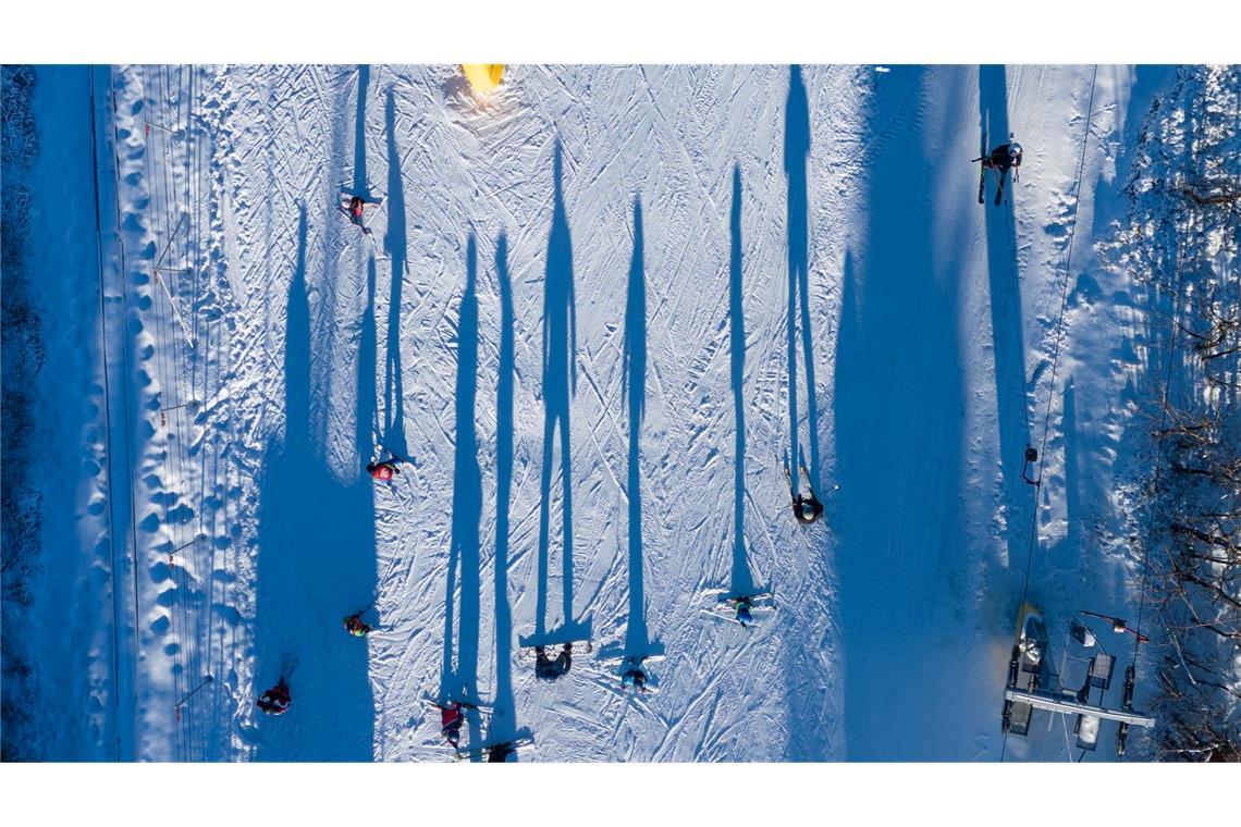 Die Aufnahme einer Fotodrohne zeigt aus ungewohnter Perspektive, wie Wintersportler im Skigebiet Erlebnisberg Altenberg neben einem Skilift die Piste hinunterfahren.