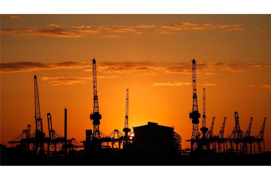 Die Sonne geht hinter Containerbrücken und Hafenkränen im Hamburger Hafen unter.