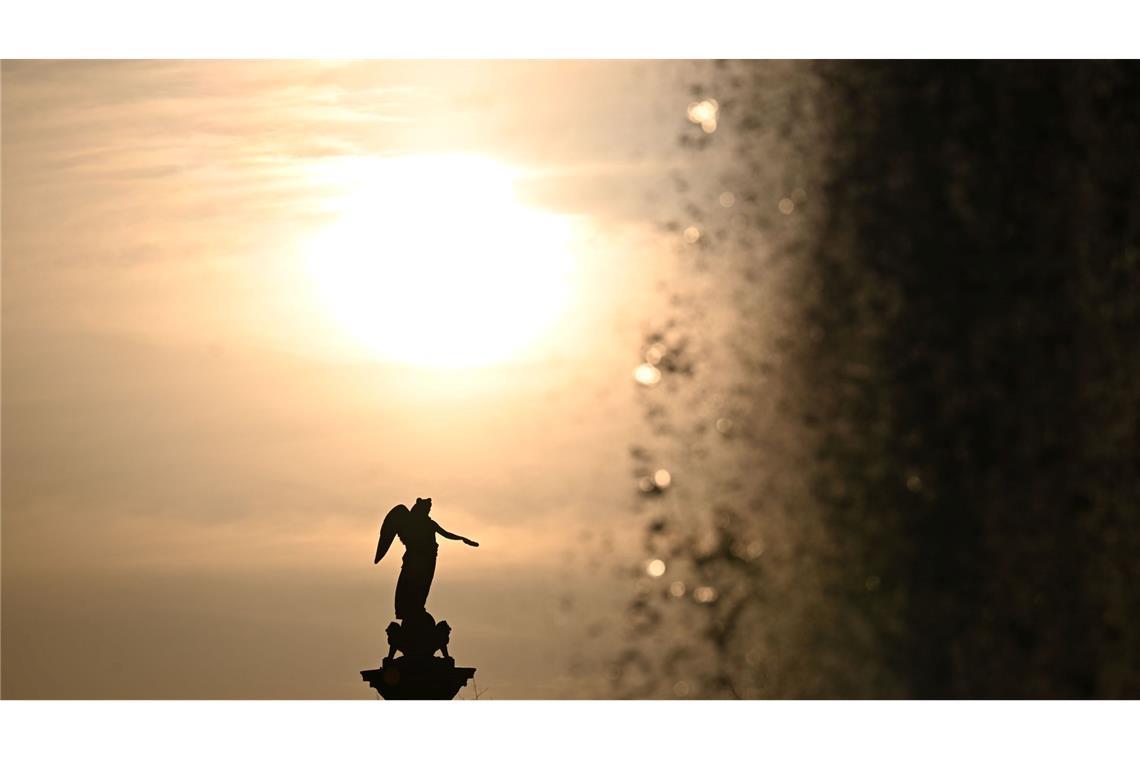 Die Sonne geht vor der Jubiläumssäule auf dem Schlossplatz unter, rechts die Fontäne eines Wasserspiels.