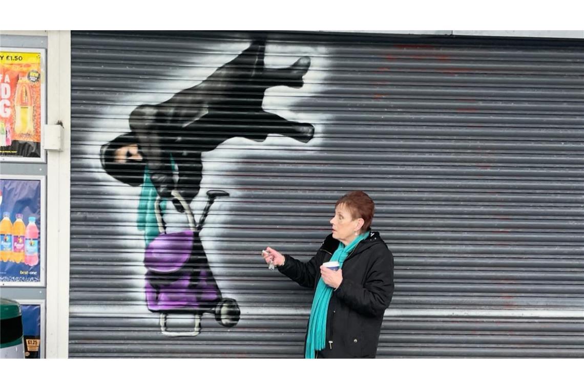 Dieses von Tee2Sugars herausgegebene Foto zeigt Anne Hughes mit dem Kunstwerk von Tee2Sugars, das sie vor dem Best One Convenience Store zeigt, wie sie vom Boden abhebt. Der lokale Künstler in Wales hat die Frau gewürdigt, die das Internet im Sturm eroberte, als sie sich im beweglichen Rollladen eines Ladens verfangen hatte.