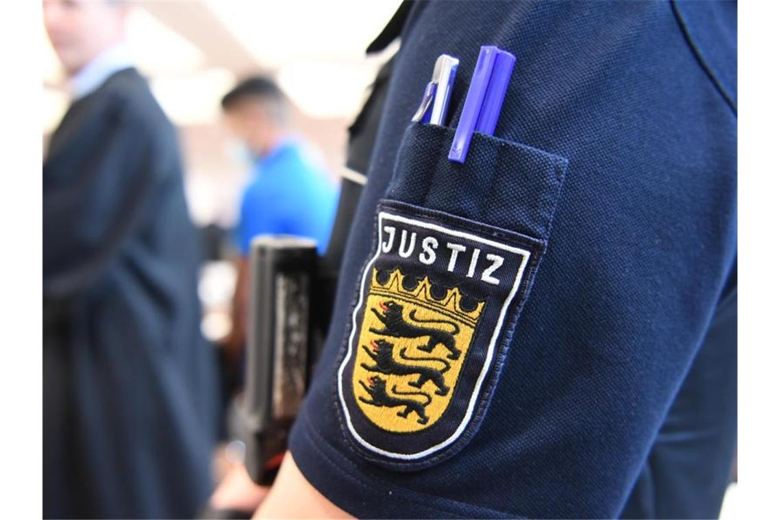 Haftstrafen gegen vier Männer wegen Vergewaltigung Ein Angeklagter steht im Gerichtssaal in der Messe Ulm, während im Vordergrund eine Justizmitarbeiterin steht, deren Justiz-Logo zu sehen ist. Foto: Felix Kästle/dpa/Archivbild
