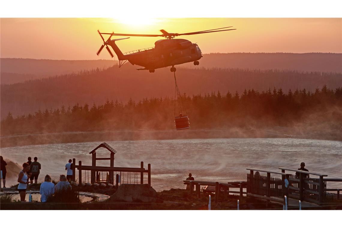 Ein Hubschrauber der Bundeswehr fliegt im Licht der untergehenden Sonne mit einem Wasserbehälter zum Königsberg am Brocken, um dort beim Kampf gegen den großen Waldbrand zu helfen.