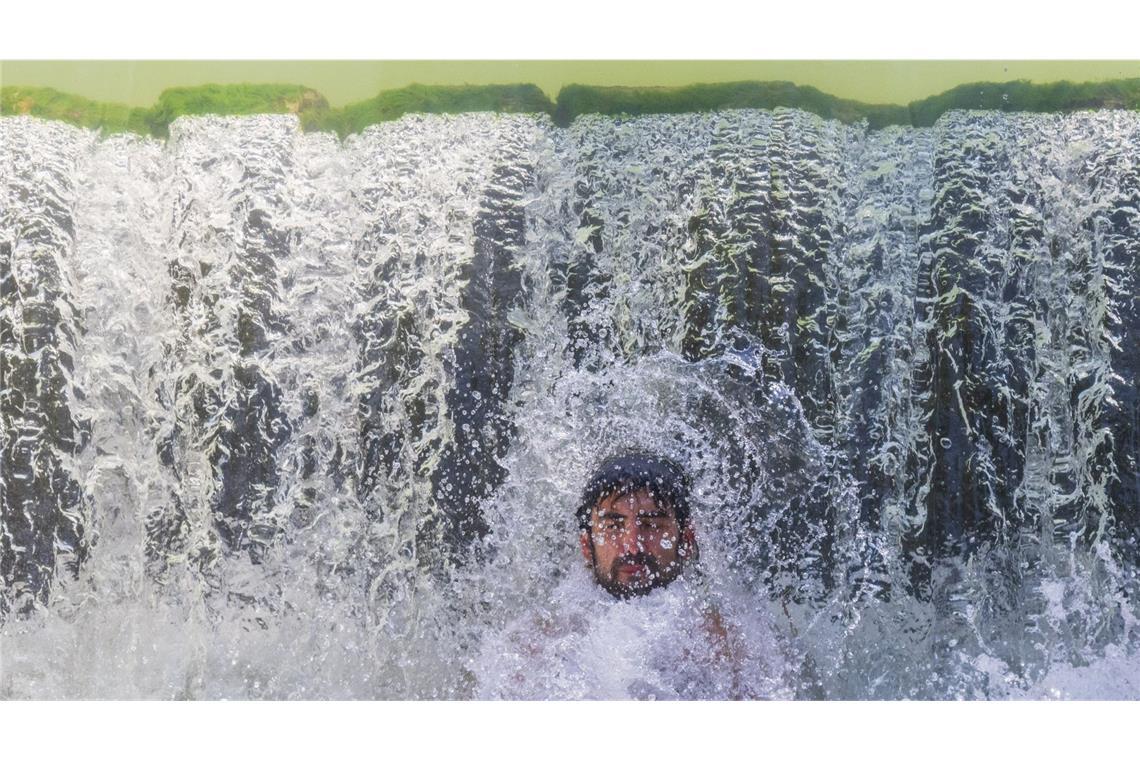 Ein junger Mann erfrischt sich am 21. Juli 2024 im Eisbach im Englischen Garten in München an einem Wasserfall.