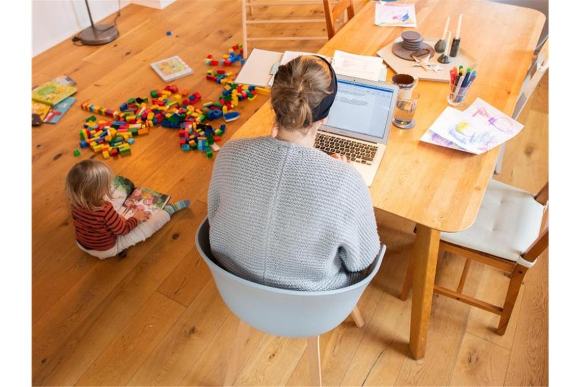 Ein Kind spielt im Wohnzimmer, während seine Mutter im Homeoffice an einem Laptop arbeitet. Foto: Julian Stratenschulte/dpa/Illustration