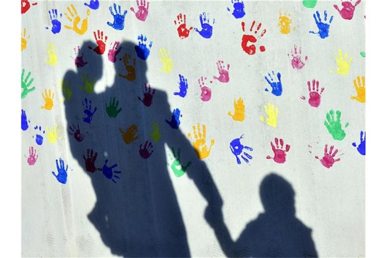 Ein Mann mit einem Kind auf dem Arm und einem an der Hand wirft einen Schatten auf eine mit bunten Handabdrücken bemalte Wand einer Kindertagesstätte. Foto: Peter Kneffel/dpa/Archiv