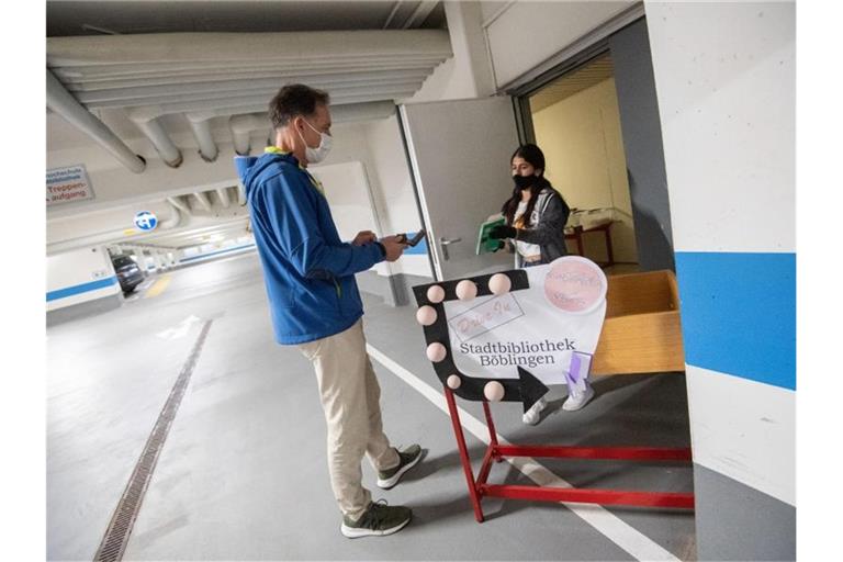 Ein Mitarbeiterin der Stadtbibliothek (r) bringt in einer Tiefgarage an einem "Drive-In"-Schalter einem Kunden ein vorbestelltes Buch. Foto: Marijan Murat/dpa