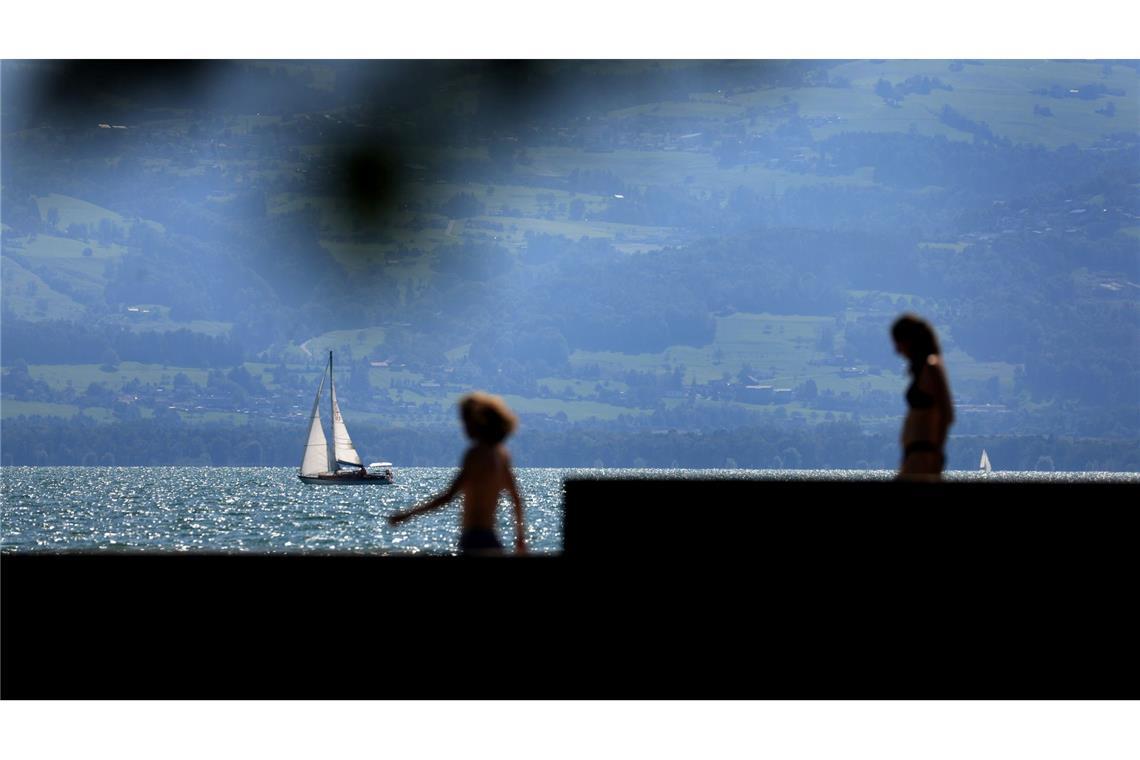 Ein Segelboot fährt hinter Badegästen auf dem im Sonnenschein glitzernden Bodensee.