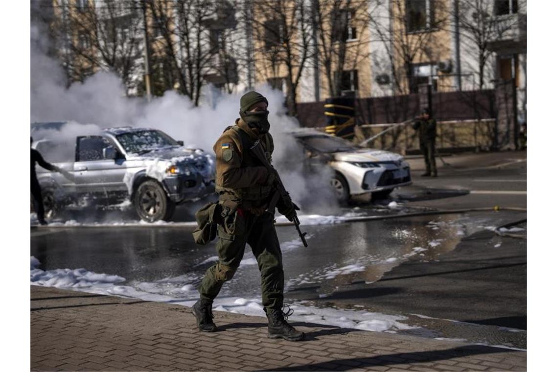 Ein ukrainischer Soldat bezieht Stellung vor einer Militäreinrichtung, während Feuerwehrleute im Hintergrund zwei brennende Autos löschen. Foto: Emilio Morenatti/AP/dpa