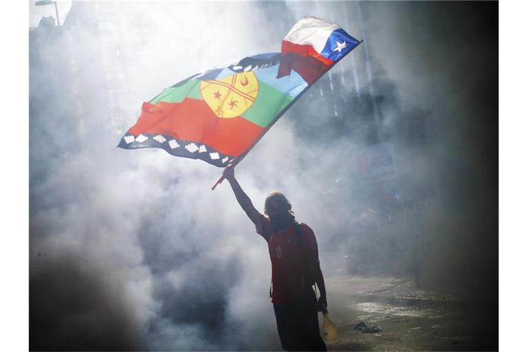 Ein vermummter Demonstrant weht die Flagge von Chile und der indigenen Gemeinschaft Mapuche mitten im Rauch während eines Protestes für Sozialreformen. Foto: Agenciauno/Agencia Uno/dpa