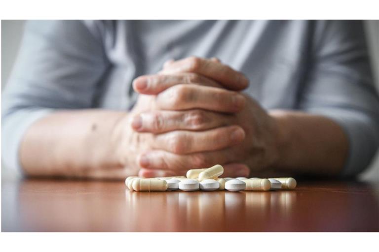 Eine Frau sitzt mit gefalteten Händen an einem Tisch. Vor ihr liegt ein Haufen Tabletten (gestellte Szene).