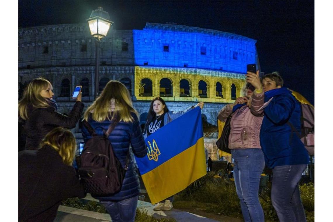 In Italien lebende ukrainische Staatsangehörige stehen vor dem Kolosseum, das in den Farben der ukrainischen Nationalflagge beleuchtet ist. Foto: Domenico Stinellis/AP/dpa