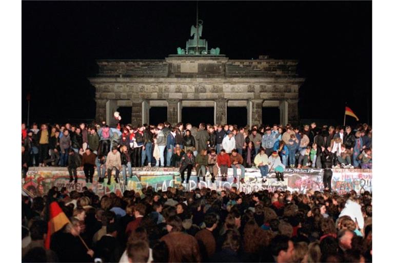 Jubelnde Menschen sitzen nach der Maueröffnung auf den Grenzanlagen am Brandenburger Tor. Foto: Wolfgang Kumm/dpa