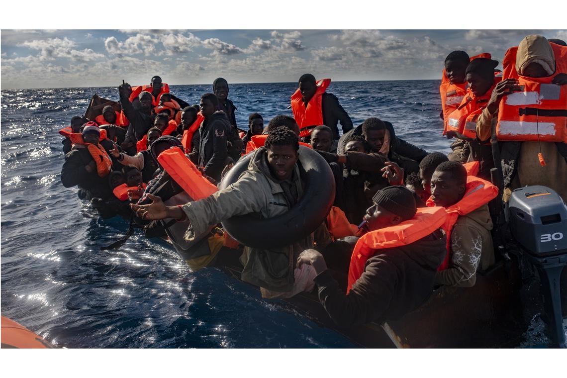 Mehrere Migranten sitzen in einem Boot im Mittelmeer, während Rettungskräfte versuchen ihnen zu helfen.