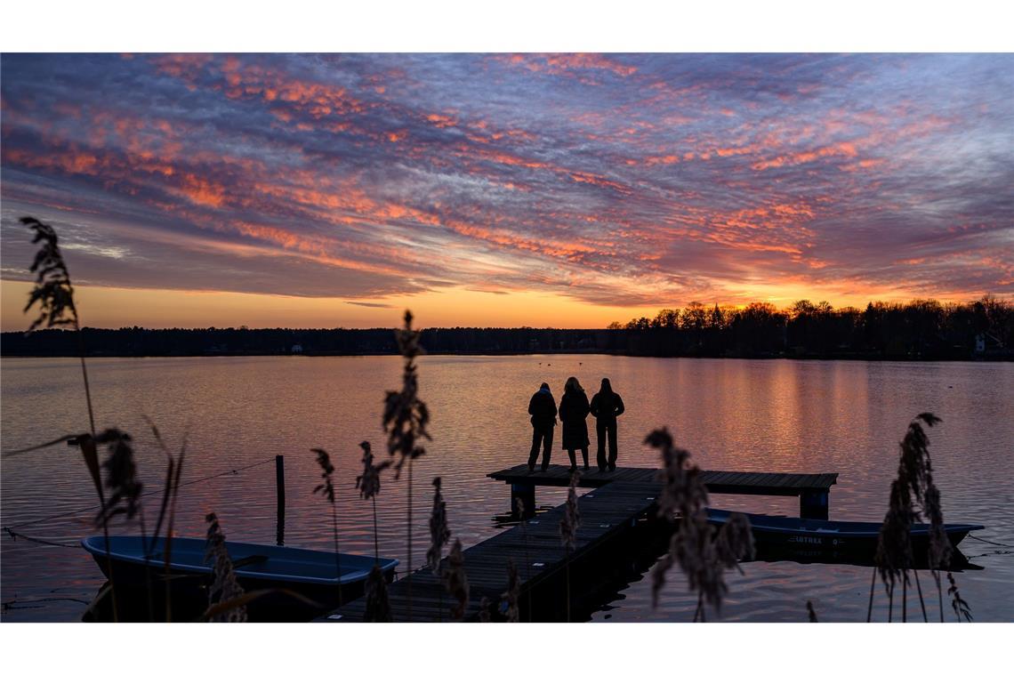 Menschen auf einem Steg beobachten, wie die Wolken zum Sonnenuntergang über dem Großen Müllroser See im östlichen Brandenburg farbenprächtig leuchten.