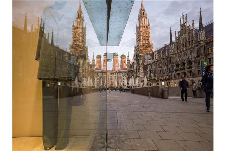 Passanten gehen über den Marienplatz in der Münchner Innenstadt. Hintergrund sind die Zwillingstürmen der Frauenkirche (l) und das Neue Rathaus (r) zu sehen. Foto: Peter Kneffel/dpa