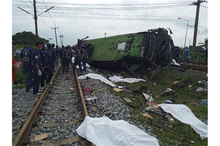 Rettungskräfte stehen an einem beschädigten Zug und neben mit weißen Tüchern bedeckten Leichen. Mindestens 17 Menschen sind am Sonntag in Thailand beim Zusammenstoß eines Busses mit einem Zug ums Leben gekommen. Foto: Uncredited/Daily News/AP/dpa