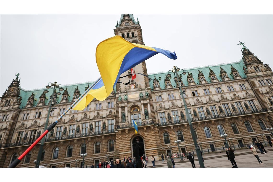 Ukraine-Krieg - Dritter Jahrestag. Ein Mann steht mit ukrainischer Fahne und Trauerflor bei der Gedenkminute „Hamburg steht still“ vor dem Rathaus.